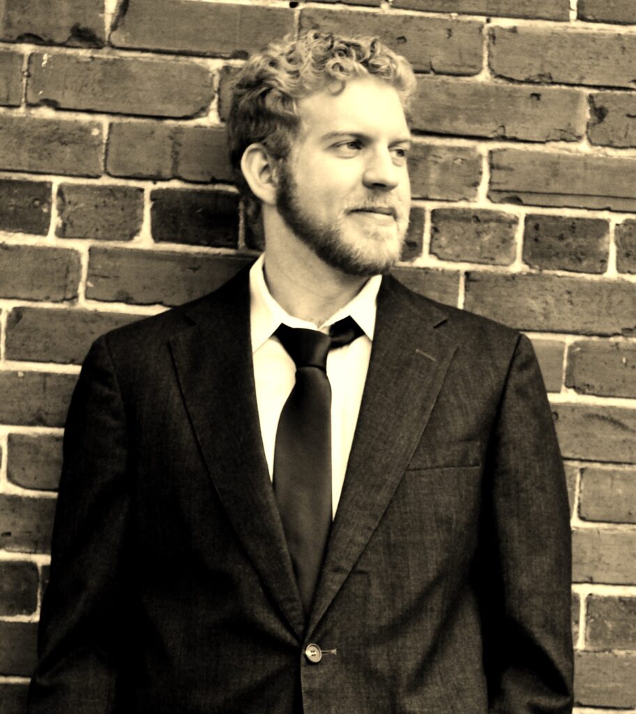 Photo of Jon Tucker in front of a brick wall looking right. Photo is in a sepia tone.
